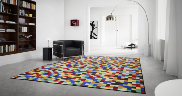 „Modernes Wohnzimmer mit farbenfrohem Vorwerk Teppich im Pixelmuster aus roten, blauen, gelben, grünen, schwarzen und weißen Quadraten; umgeben von minimalistischer Einrichtung mit schwarzem Sessel, weißer Lampe und Bücherregal.“